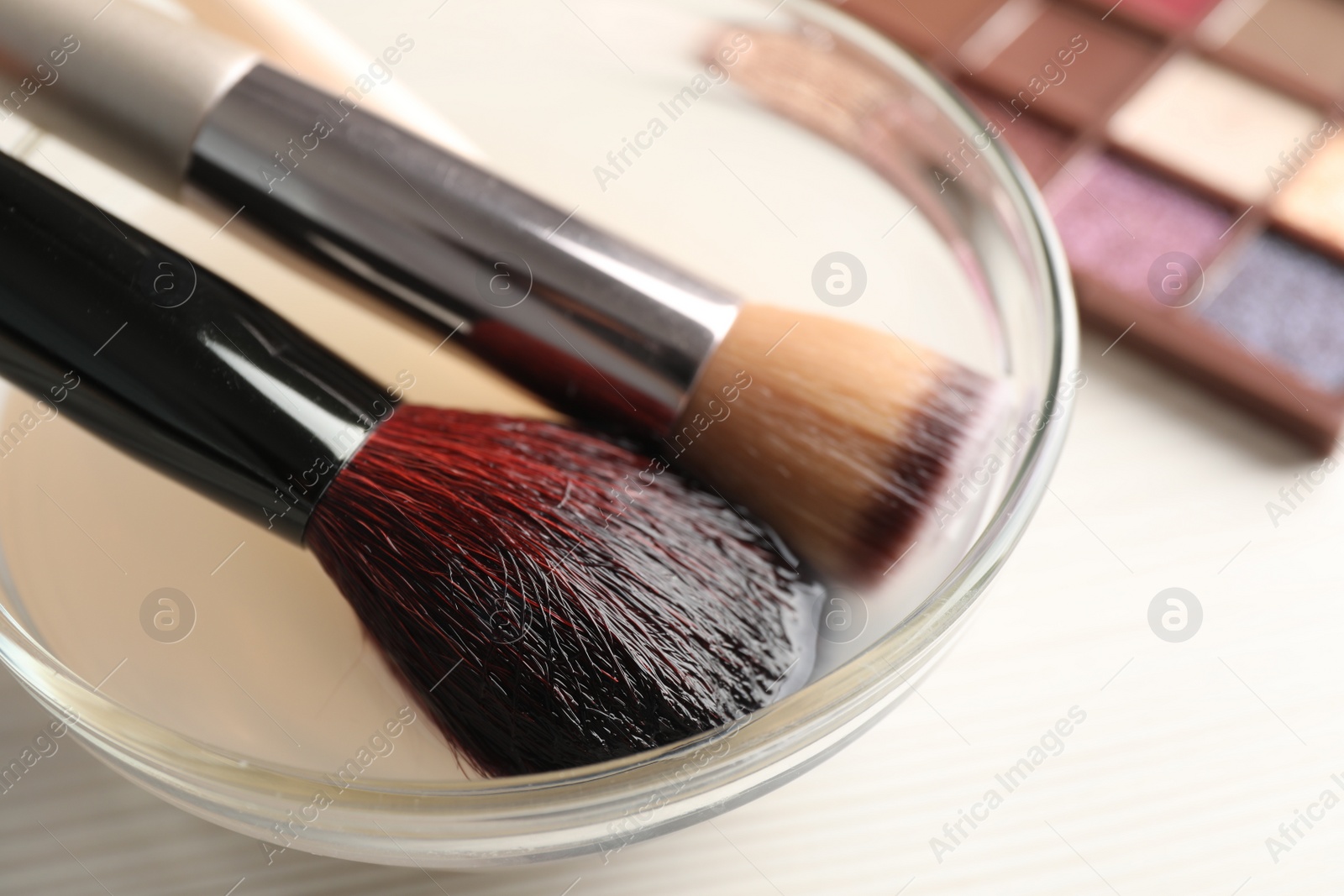 Photo of Cleaning makeup brushes in bowl with special liquid on white wooden table, closeup