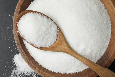 Granulated sugar in bowl and spoon on black table, top view