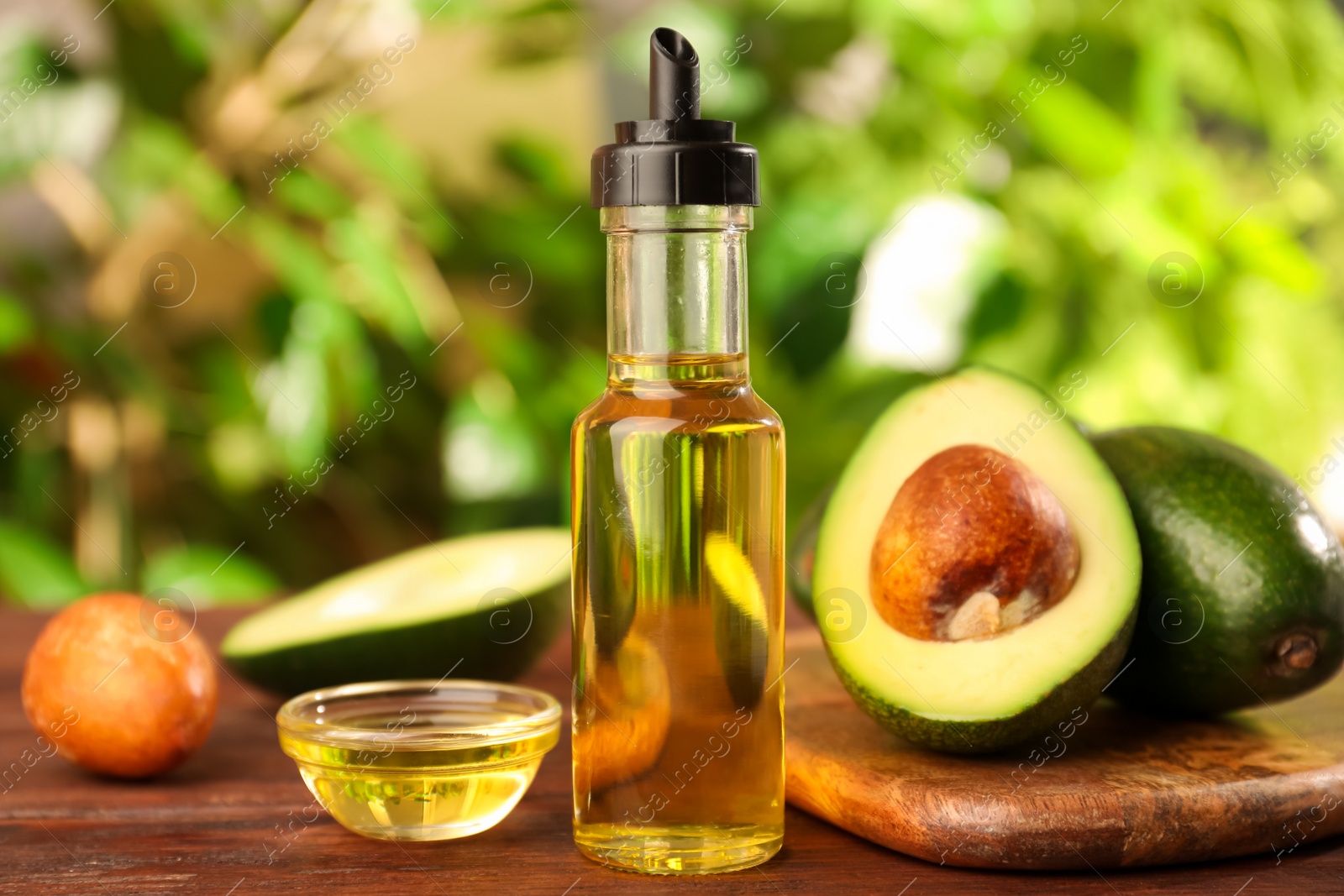 Photo of Glass bottle of cooking oil and fresh avocados on wooden table against blurred green background