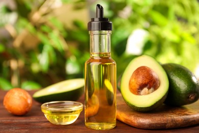 Photo of Glass bottle of cooking oil and fresh avocados on wooden table against blurred green background