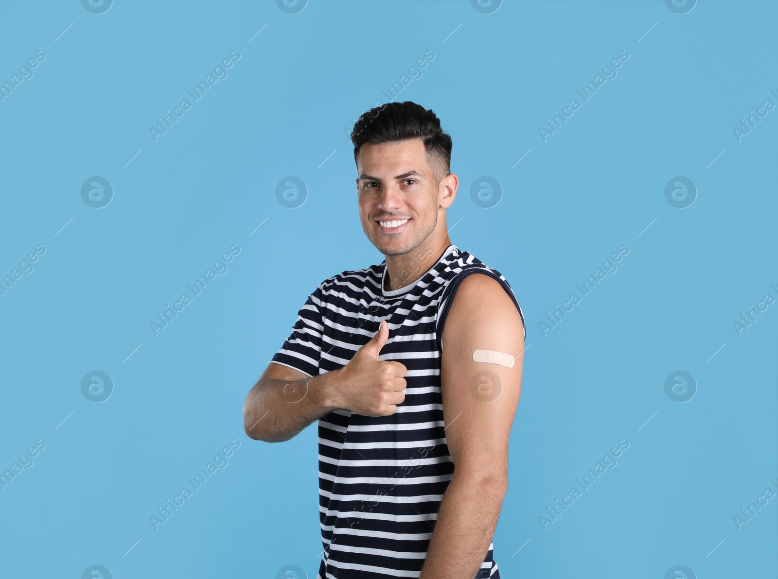 Photo of Vaccinated man with medical plaster on his arm showing thumb up against light blue background