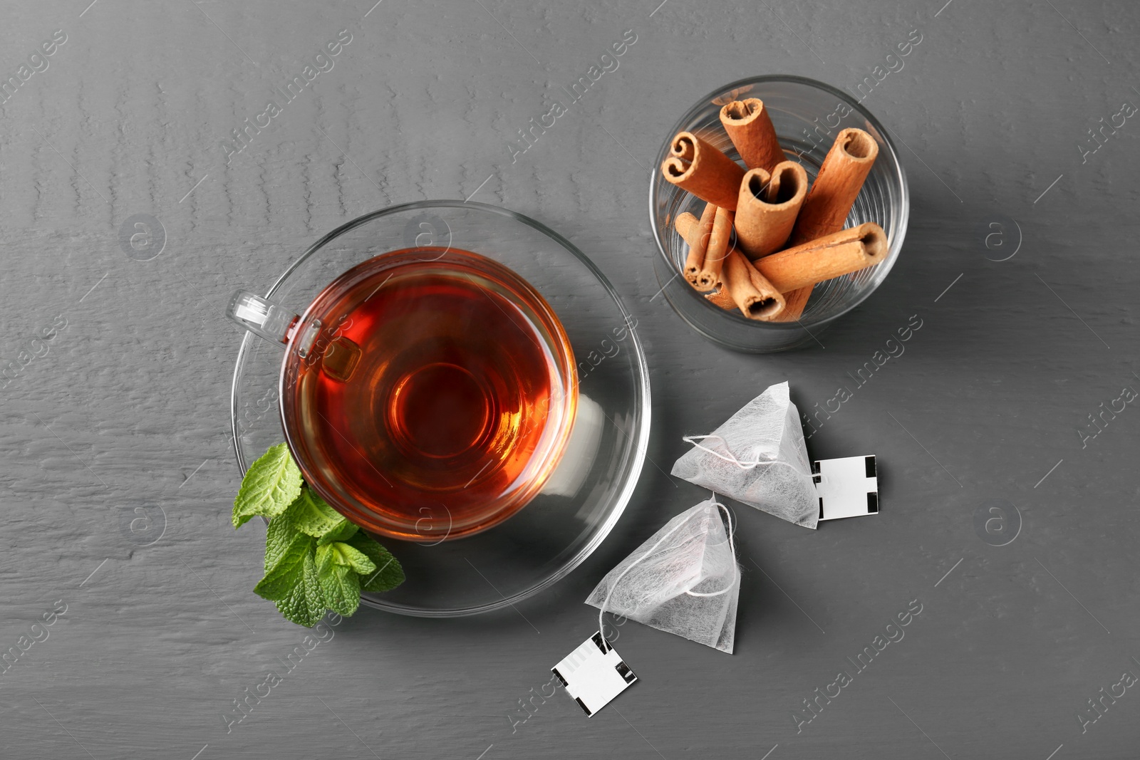 Photo of Tea bags, cup of hot drink, mint and cinnamon sticks on grey wooden table, flat lay