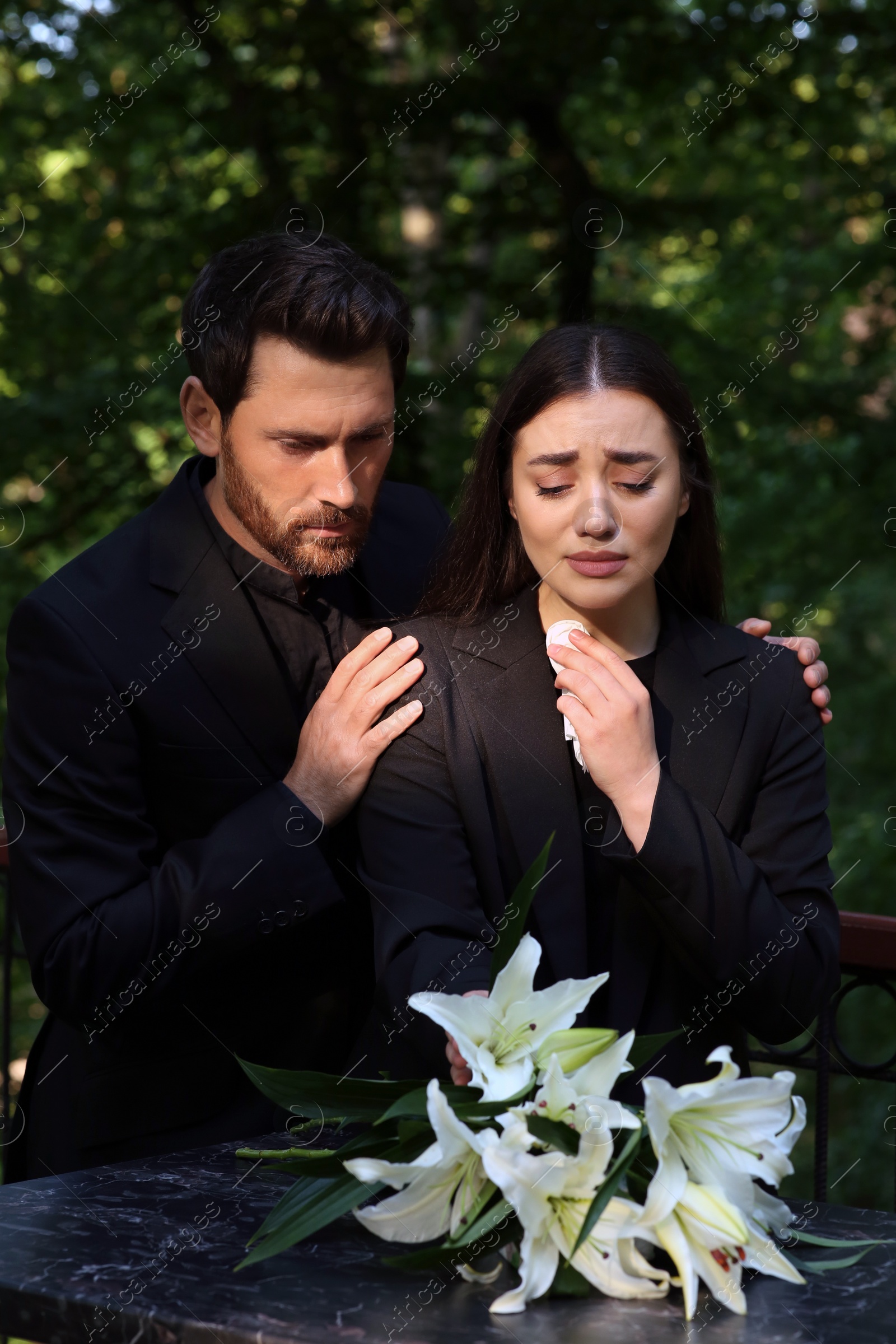 Photo of Sad couple mourning near granite tombstone with white lilies at cemetery. Funeral ceremony