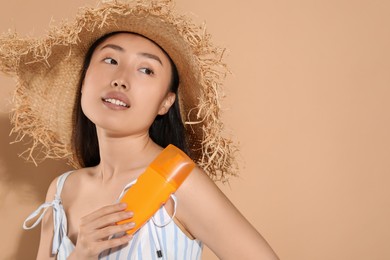 Beautiful young woman in straw hat holding sun protection cream on beige background, space for text