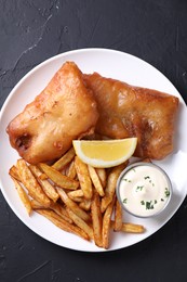 Photo of Tasty fish, chips, lemon and sauce on black table, top view