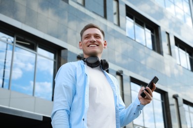 Smiling man with headphones and smartphone near building outdoors, low angle view