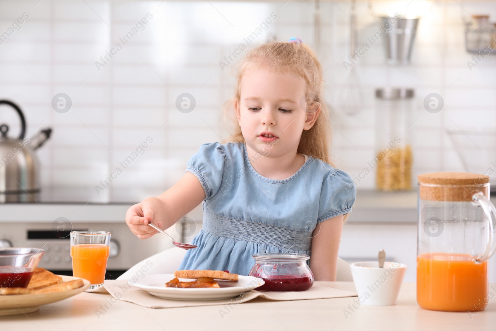 Photo of Cute little girl spreading jam onto tasty toasted bread at table in kitchen