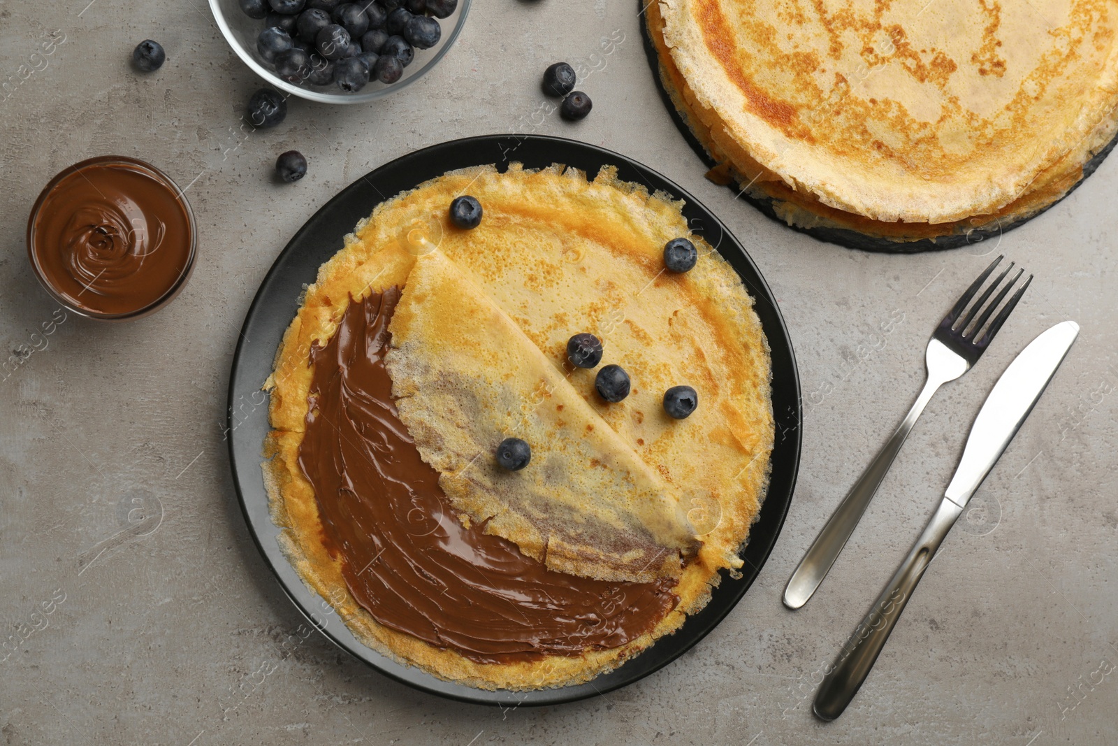 Photo of Delicious thin pancakes with chocolate spread and blueberry on grey table, flat lay