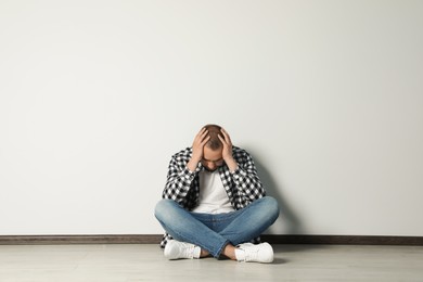 Sad young man sitting on floor near white wall indoors, space for text