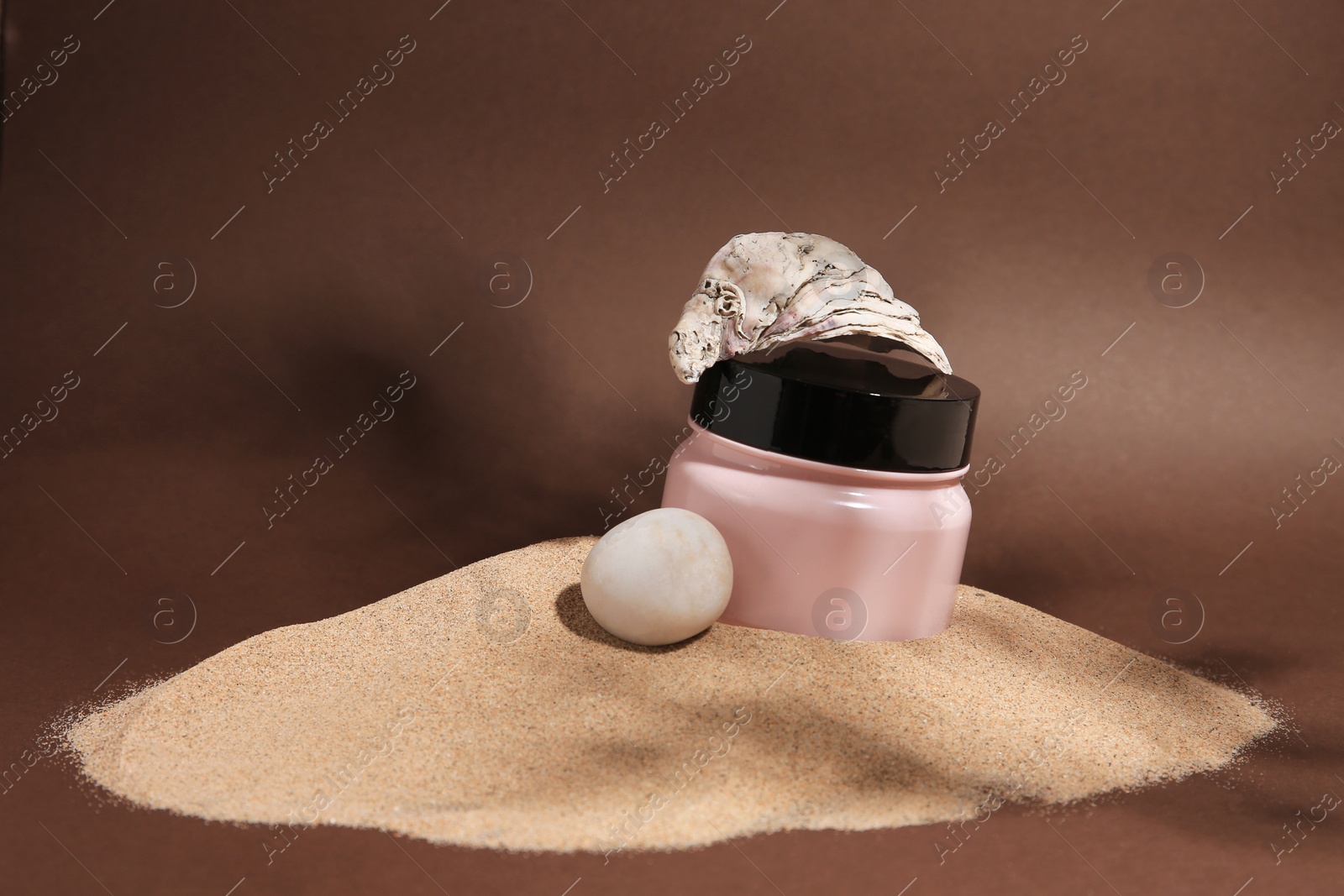 Photo of Jar of body cream, seashell and stone on sand against brown background. Space for text