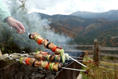 Photo of Woman cooking meat and vegetables on brazier against mountain landscape, closeup