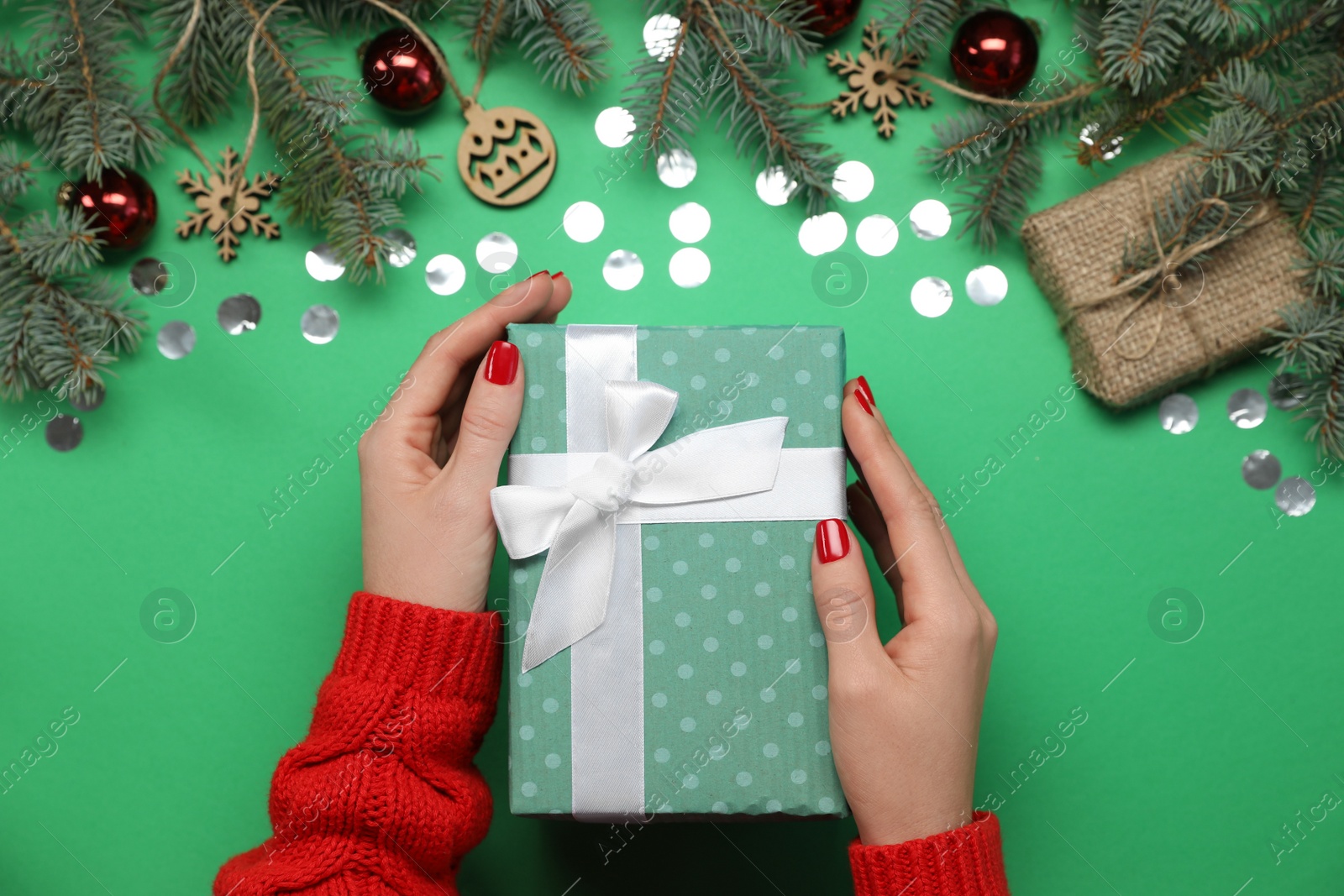 Photo of Woman holding gift box near fir tree branches with Christmas decor on green background, top view