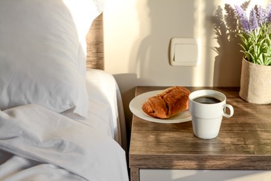 Photo of Cup of morning coffee and croissant on wooden night stand near bed indoors