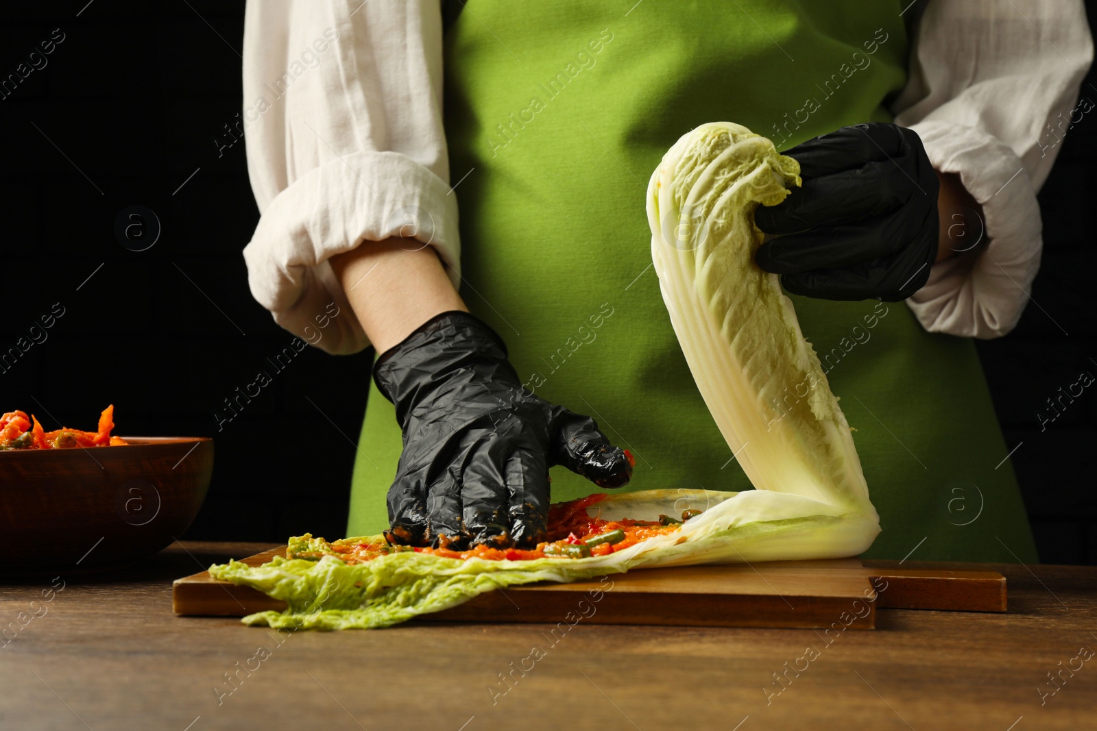 Photo of Woman preparing spicy cabbage kimchi at wooden table against dark background, closeup