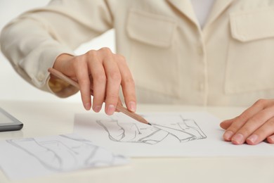Photo of Woman drawing clothes with pencil on sheet of paper at white table, closeup