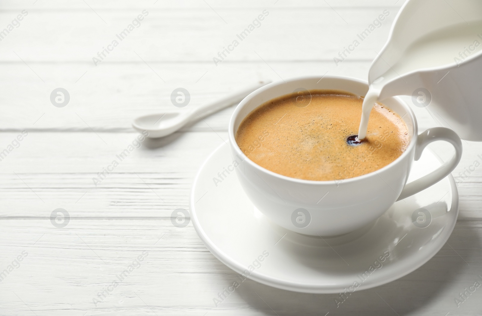 Photo of Pouring milk into cup of hot coffee on white wooden table, space for text