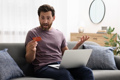 Photo of Shocked man with credit card and laptop on sofa at home. Be careful - fraud