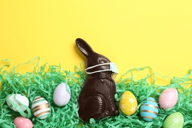 Photo of Chocolate bunny with protective mask and eggs on yellow background, flat lay. Easter holiday during COVID-19 quarantine