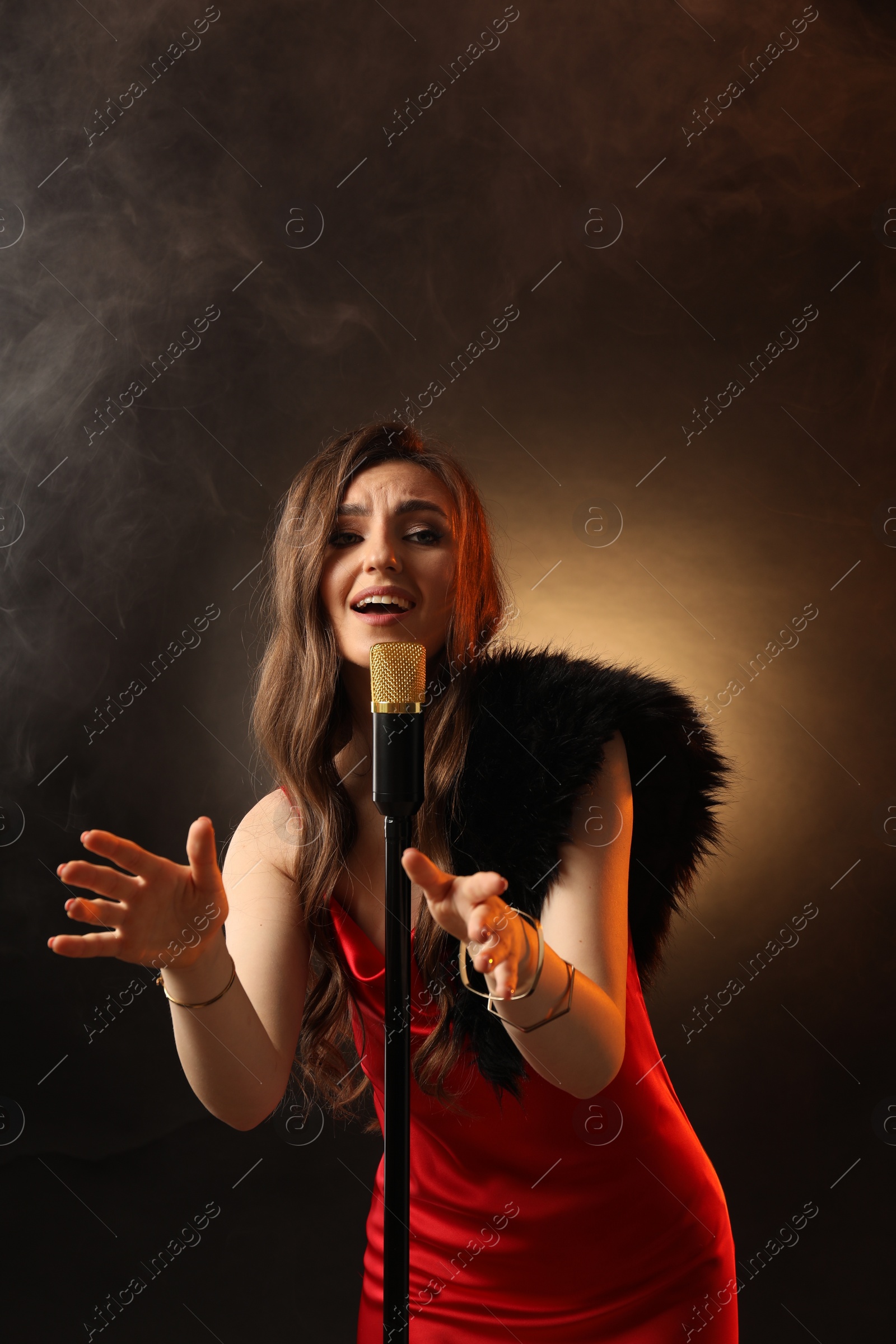 Photo of Beautiful young woman in stylish dress with microphone singing on dark background in color lights and smoke