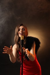 Beautiful young woman in stylish dress with microphone singing on dark background in color lights and smoke