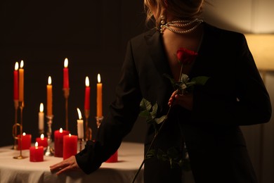 Photo of Woman holding beautiful rose near table with burning candles at night, back view