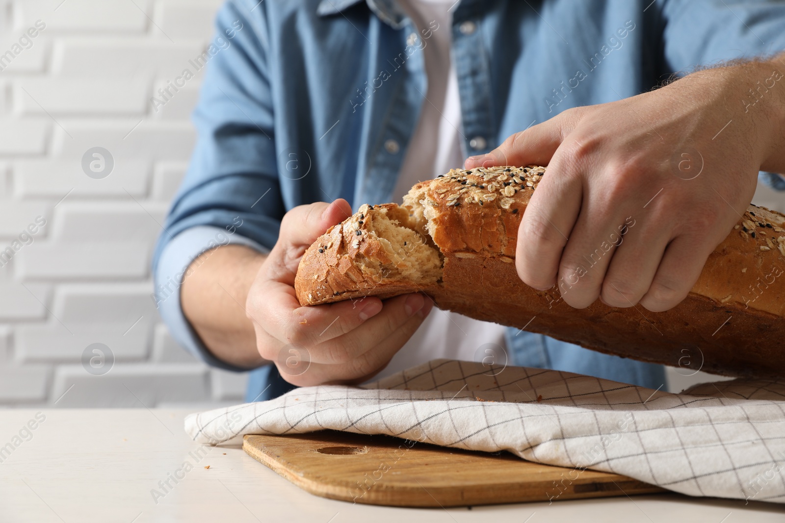 Photo of Man breaking loaf of fresh bread at white table near brick wall, closeup