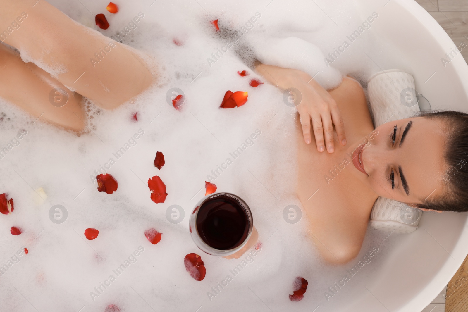 Photo of Woman with glass of wine taking bath in tub with foam and rose petals, top view