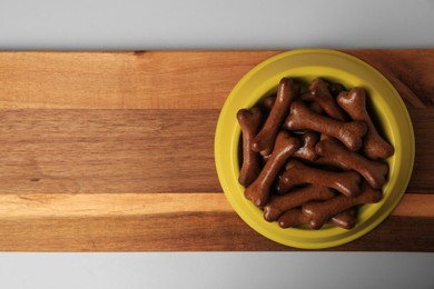 Photo of Yellow bowl with bone shaped dog cookies on wooden board, top view. Space for text