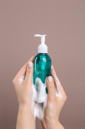 Photo of Woman with foam on hands holding bottle of skin cleanser against brown background, closeup. Cosmetic product