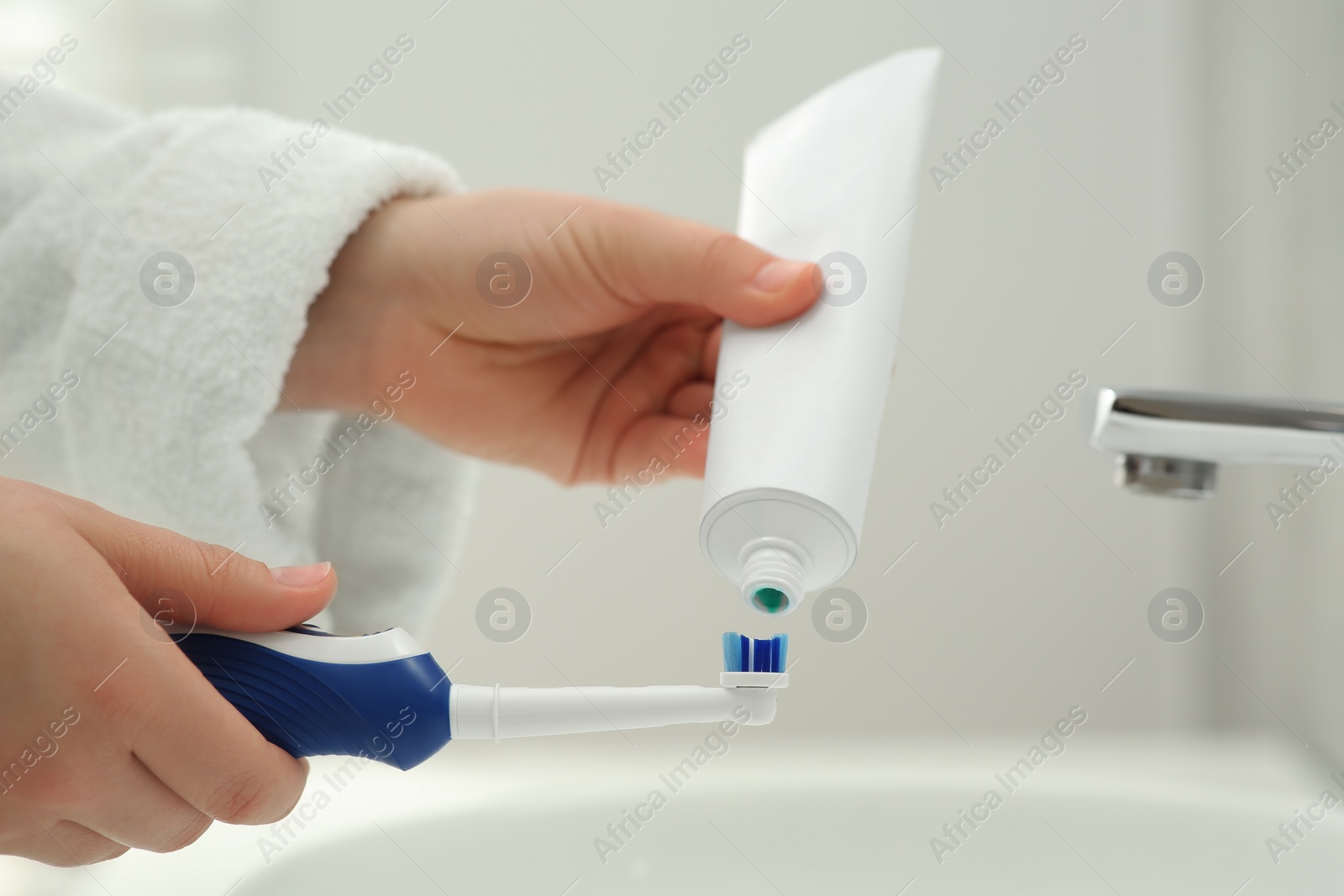 Photo of Woman squeezing toothpaste from tube onto electric toothbrush above sink in bathroom, closeup