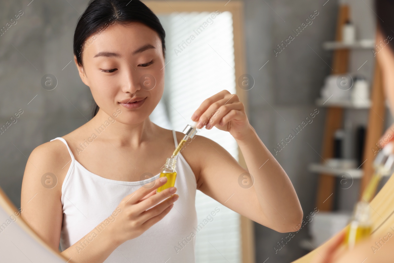 Photo of Beautiful young woman with bottle of cosmetic serum near mirror in bathroom