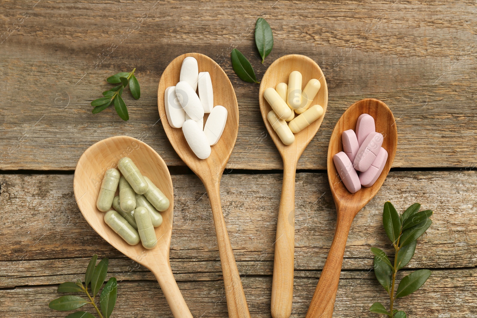 Photo of Vitamin capsules in spoons and leaves on wooden table, flat lay