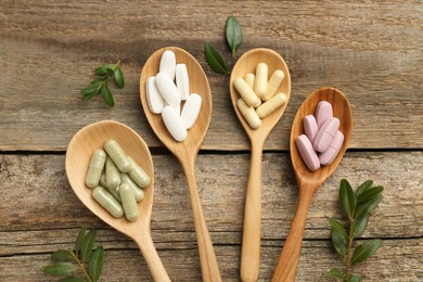 Photo of Vitamin capsules in spoons and leaves on wooden table, flat lay