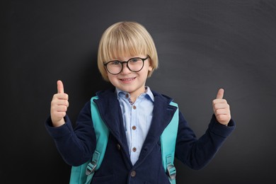 Photo of Happy little school child with backpack showing thumbs up near chalkboard