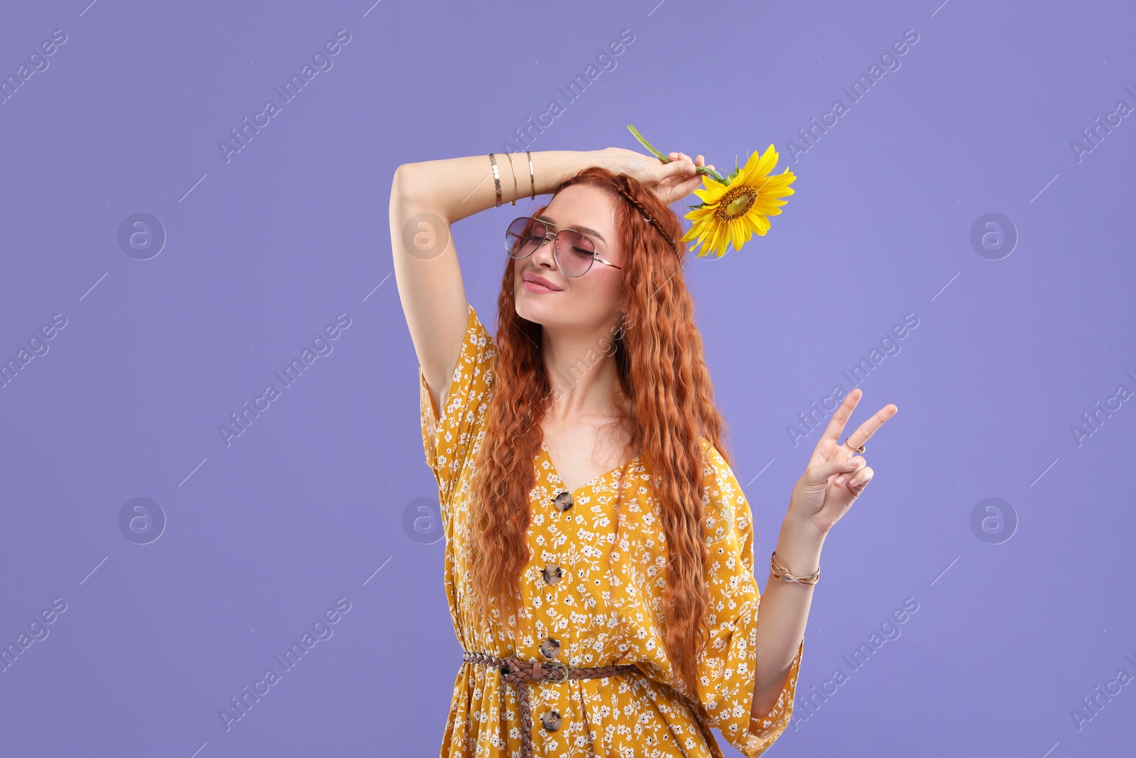 Photo of Beautiful young hippie woman with sunflower showing V-sign on violet background