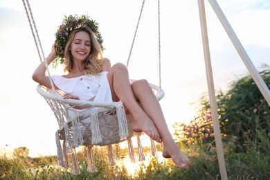 Photo of Young woman wearing wreath made of beautiful flowers on swing chair outdoors at sunset