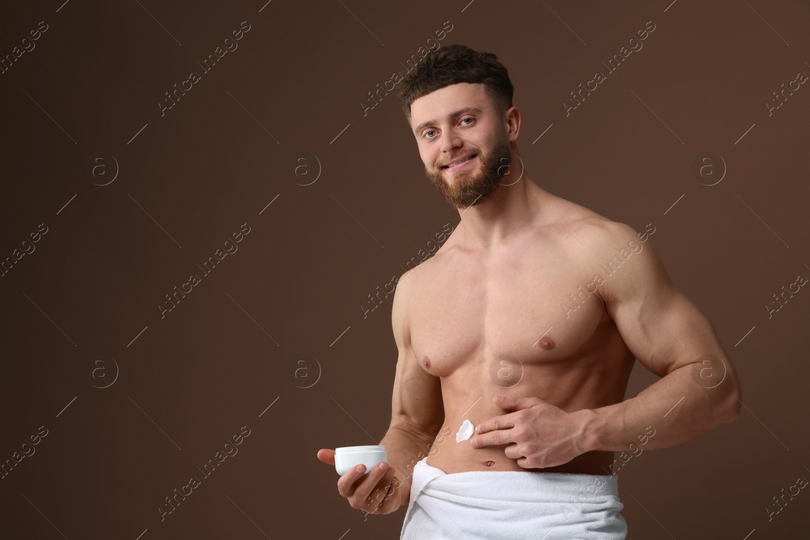 Photo of Handsome man applying moisturizing cream onto his body on brown background, space for text