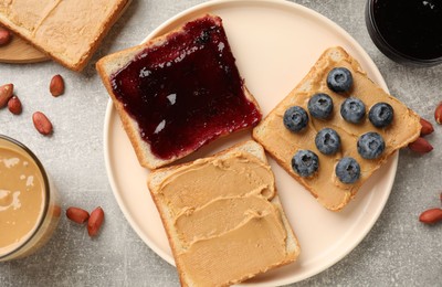 Photo of Tasty peanut butter sandwiches with jam, fresh blueberries and peanuts on gray table, flat lay