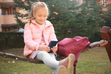 Cute little girl with backpack and smartphone on wooden swing outdoors
