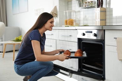 Photo of Beautiful woman taking out tray of baked buns from oven in kitchen