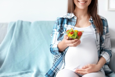 Young pregnant woman with vegetable salad on sofa at home