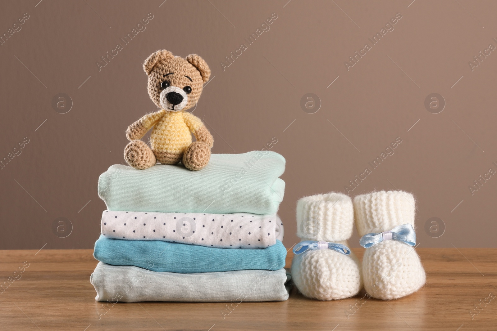 Photo of Stack of baby boy's clothes, booties and toy on wooden table against brown background, space for text