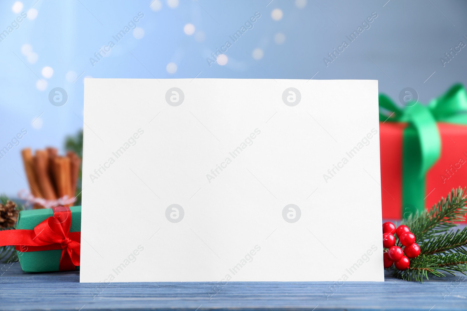 Photo of Blank Christmas card and festive decor on blue wooden table against blurred lights, closeup. Space for text