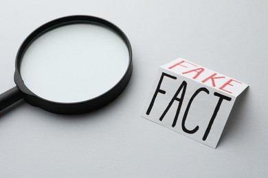 Photo of Magnifying glass near sheet of paper with words Fact and Fake on light grey background