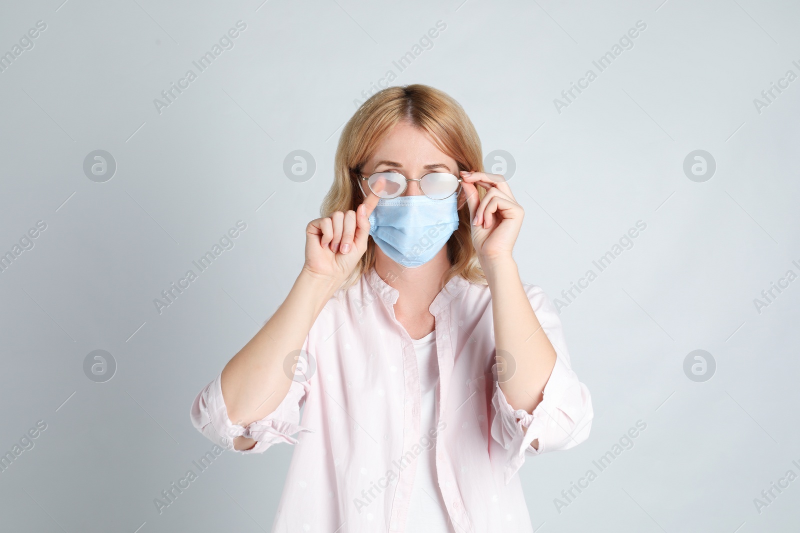 Photo of Woman wiping foggy glasses caused by wearing medical mask on light background