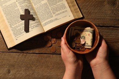 Donate and give concept. Woman holding bowl with money. Bible and cross on wooden table, top view