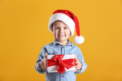 Photo of Cute little boy in Santa Claus hat holding gift box on yellow background