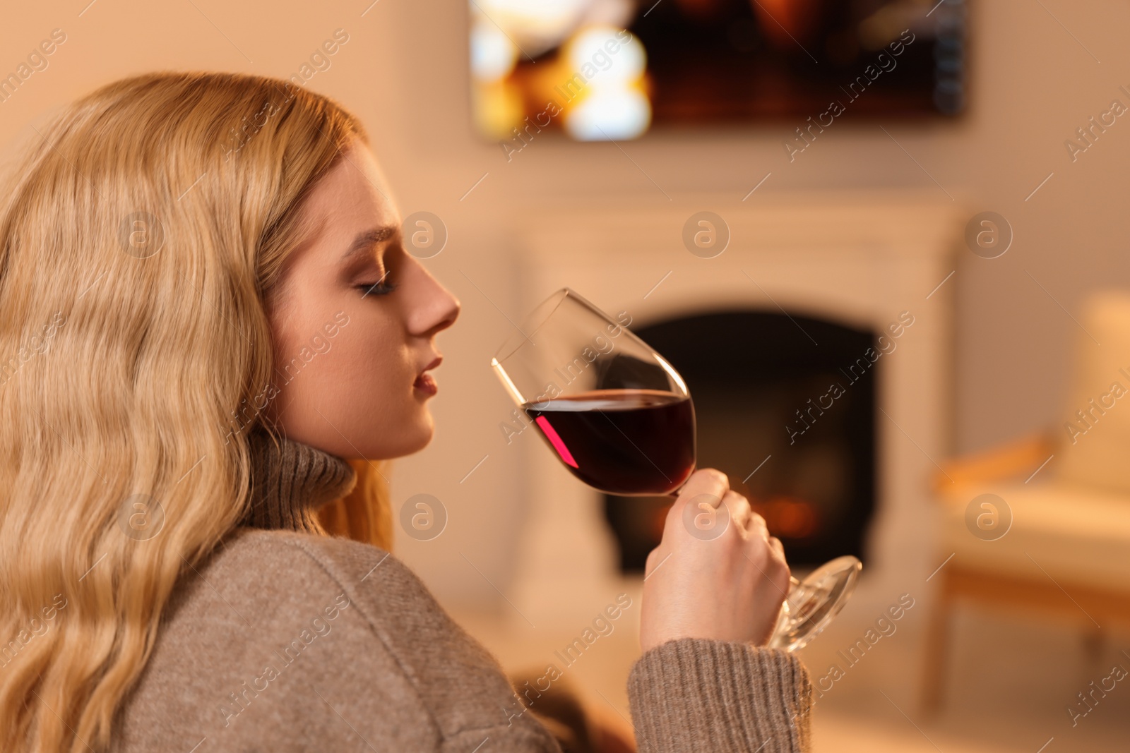 Photo of Beautiful young woman with glass of wine resting near fireplace at home