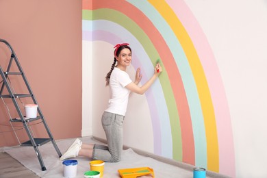 Young woman painting rainbow on white wall indoors