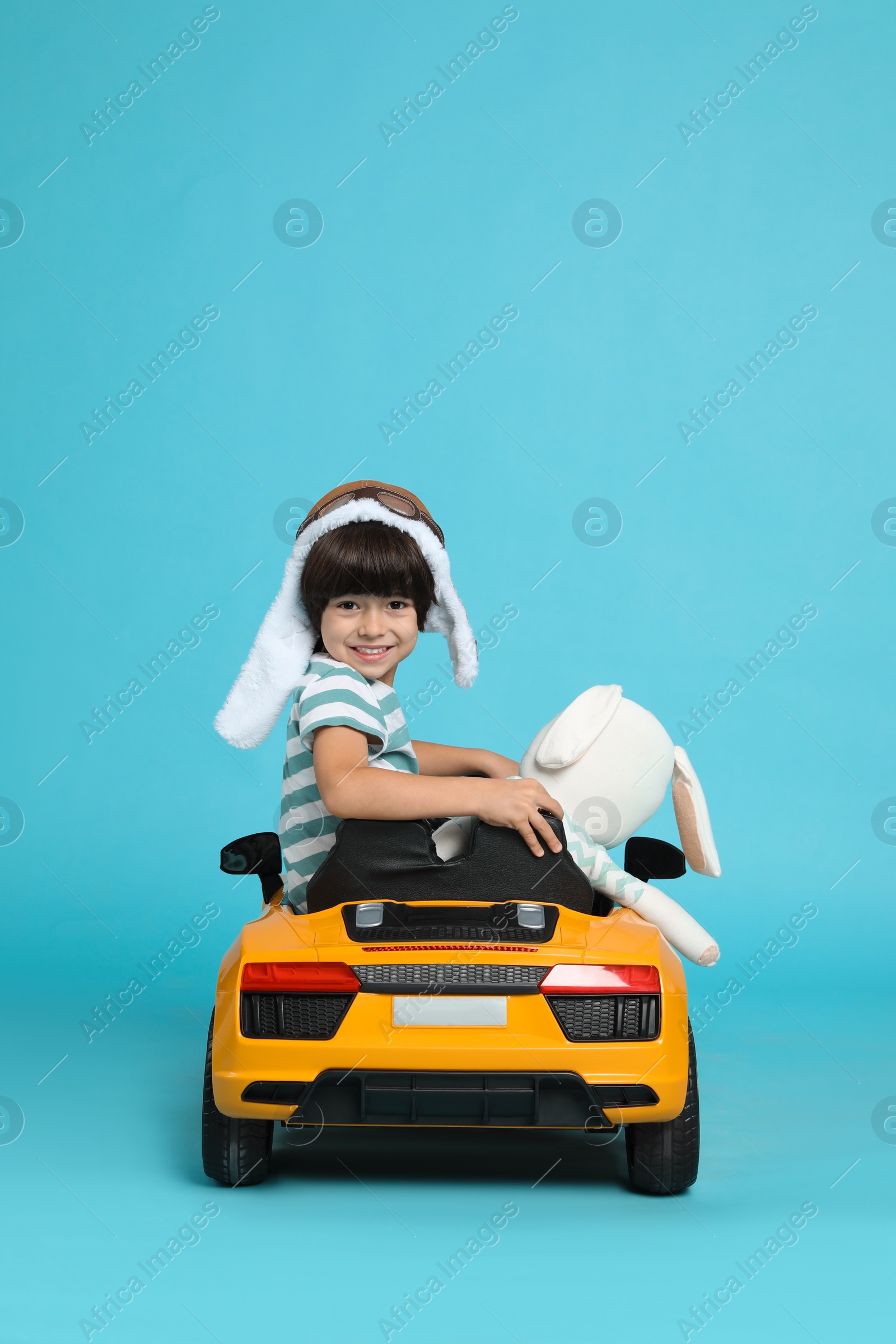 Photo of Cute little boy with toy bunny driving children's car on light blue background